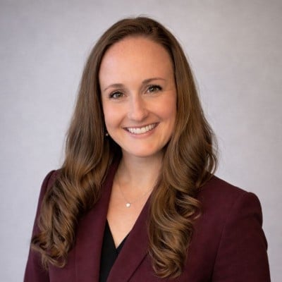Professional headshot of a smiling woman with long brown hair wearing a maroon blazer over a black top